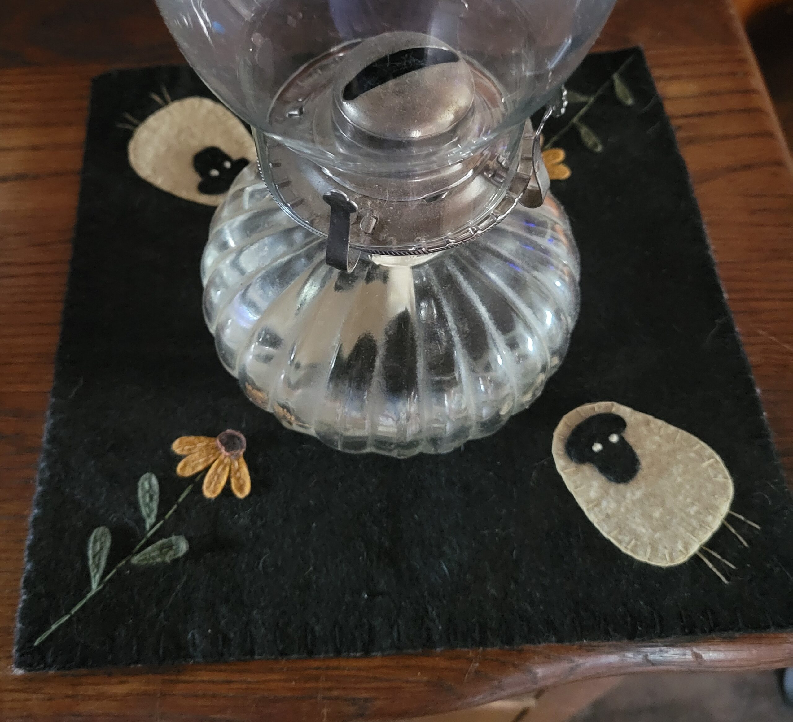 glass with square mat of gray felt with embroidered felted sheep and flowers in corners underneath